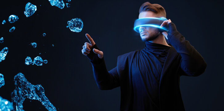 Beautiful young man using VR helmet while touching air in blue neon lights. Guy in glasses of virtual reality with blue backlight over dark magic background. 