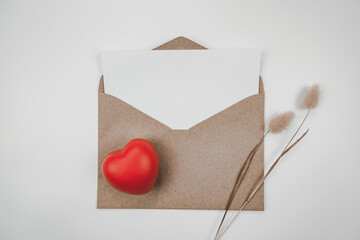 Blank white paper is placed on open brown paper envelope with red heart with Rabbit tail dry flower on white background. Valentine's day concept.