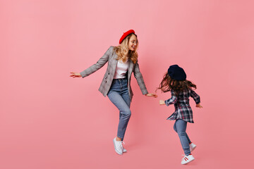 Stylish curly blond woman in jeans and gray jacket dancing with her little dark-haired daughter on pink background