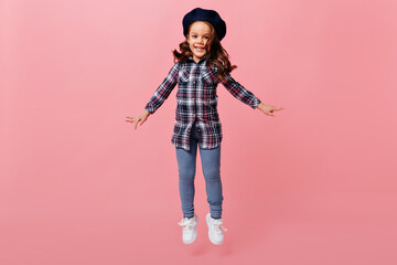 Curly positive female child with smile looks into camera and jumps. Studio photo of girl in white sneakers, jeans, plaid shirt and dark beret
