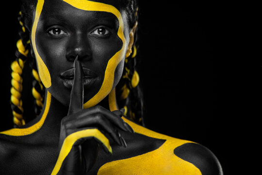 Face art. Girl showing gesture quietly, asks remain silent with finger. Woman with black and yellow body paint. Young african girl with colorful bodypaint. An amazing afro american model with makeup.