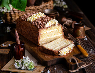 homemade rectangular layered sponge cake decorated with white and chocolate cream on rustic table with bouquet of lilac