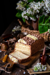 homemade rectangular layered sponge cake decorated with white and chocolate cream on rustic table with bouquet of lilac