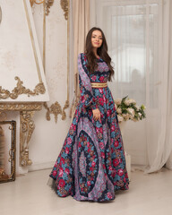 Elegant brunette young woman with long wavy hair in long colorful purple dress standing and posing in bright luxury interior