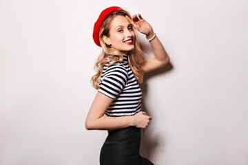 Portrait of lady with red lipstick and in bright beret smiling against white background. Girl in striped top and black skirt posing