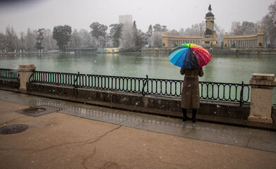 a woman with a brown jacket holding a rainbow colorful umbrella stand alone contemplating of a beautiful lake and monument with ducks in a winter cold snowfall day