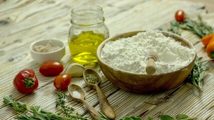 Ingredients for making Italian focaccia bread. Flour, olive oil, yeast, seasonings, salt, tomatoes, sweet herbs, greens close-up on a wooden table, copy space