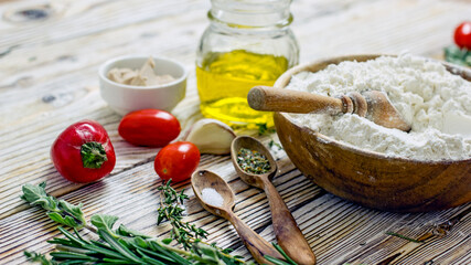 Ingredients for making Italian focaccia bread. Flour, olive oil, yeast, seasonings, salt, tomatoes, sweet herbs, greens close-up on a wooden table, copy space