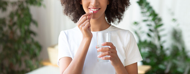 Cheerful young african american woman takes a pill of vitamins and drinks with glass of water