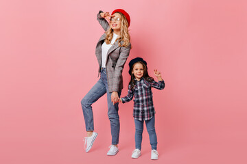 Adult woman and female child pose against pink background and show peace sign. Portrait of mother and daughter in plaid shirts and berets