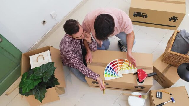 Upper view of two men sitting on the floor and looking at color pattern samples, color swatch, on a move cardboard box. Gay couple Moving into a new home concept, renovation, repairing, designing.
