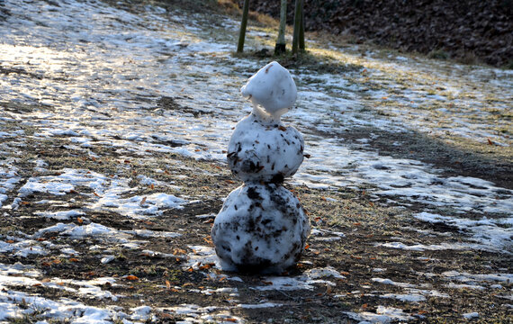 A Snowman Sadly Slowly Melting, Dirty From Mud And Lack Of Snow Will Evoke A Feeling Of Ruin And Ugly Changeable Weather Of Winter February. Frozen Snow On Lawn