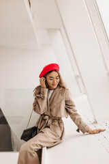 Lady in glasses and red beret, dressed in long trench coat, flirtatiously touches blonde hair, posing by window