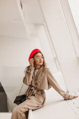 Slim lady in elegant beige trench coat and red beret touches her hair and sits by window on white background