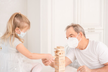 Obraz na płótnie Canvas Old grandfather wearing protective face mask plays with his little granddaughter during quarantine Coronavirus (Covid-19) epidemic