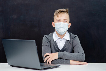 Teen schoolboy wearing protective face mask sits with laptop at school near blackboard during corona virus and flu outbreak. Empty space for text