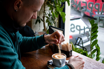 Man pouring sugar in a cup of coffee in front of a window of a cafe.
