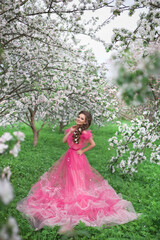 Fototapeta na wymiar A beautiful young girl with long hair in a light pink ball gown walks through a blooming apple orchard