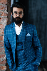 Stylish businessman stands in a suit with one hand in his pocket against the background of a window