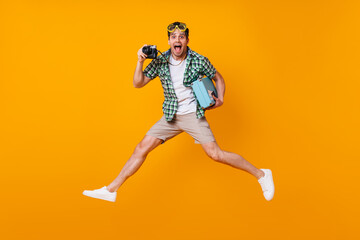 Fototapeta na wymiar Funny tourist guy in summer outfit holding retro camera and blue suitcase. Man in diving mask jumping on orange background