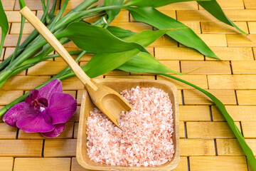 Spa, zen, massage concept. Pink himalayan bath salt with spoon, bamboo leaves, purple orchid, soap in soap dish on wooden carpet background. Top view. Close up. Selective focus. Text copy space.