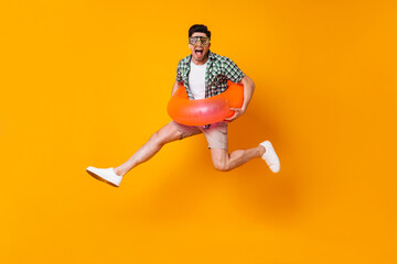Young mischievous man in summer outfit and diving mask jumping with inflatable circle on orange background