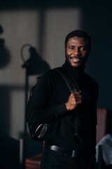A man in a black shirt stands against a dark background with a backpack on his back