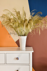 A pot with dried plants on a white dresser. Behind it, there is a wall painted with geometric shapes.