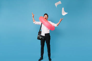 Full-length photograph of man scattering office documents on blue background. Guy in shirt and pants posing with inflatable circle