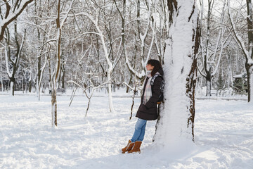 Fototapeta na wymiar Young smiling woman standing near to a big tree in a snow-covered winter park. Snowy winter