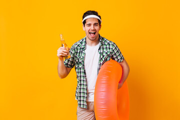 Nice guy in summer outfit winks, holds beer and rubber ring on orange background