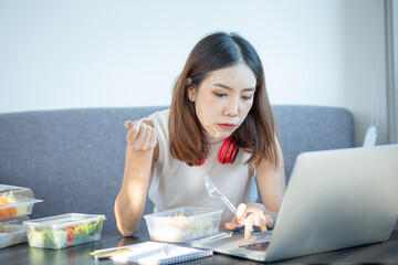 Young attractive asian female hungry eat doughnut take away snack food with full mouth look at computer notebook at home in busy work from home multitask unhealthy meal lifestyle concept.
