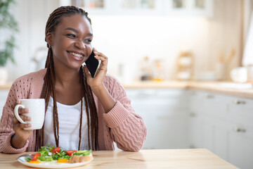 Cheerful Young African Woman Talking On Cellphone While Eating Breakfast In Kitchen
