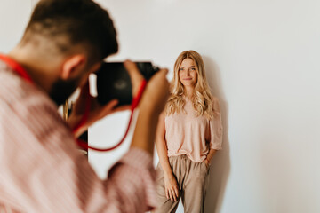 Dark-haired guy makes photo of his blond girlfriend dressed in pink T-shirt and beige pants against white wall