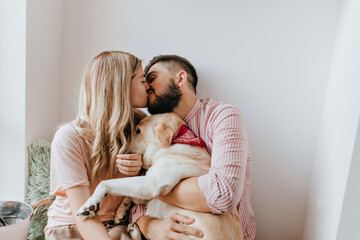 Romantic couple kisses in bright sunny room. Guy in pink shirt and his girlfriend hugging dog