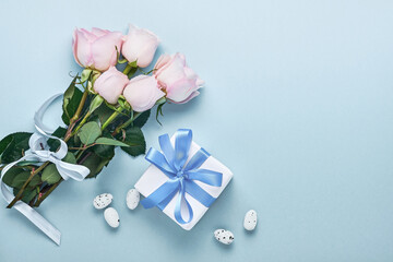 Pink roses flowers bouquet with ribbon, gift box and Easter eggs over beautiful blue background. Greeting card template with copy space