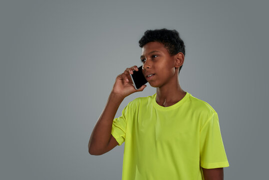 Young People And Communication Concept. Close Up Shot Of A Teenage African Boy Wearing Neon T Shirt Talking By Phone While Standing Against Grey Background