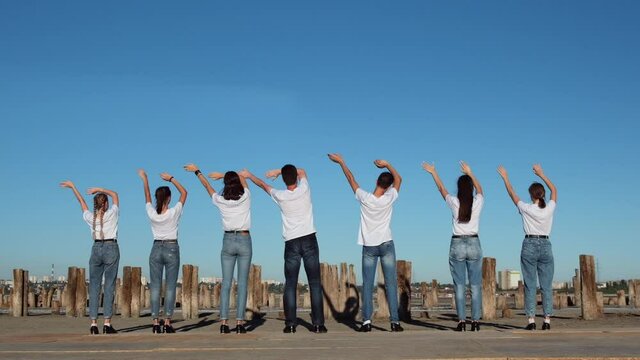 Young guys and girls in white T-shirts waving their hands in sync, standing with their backs to the video camera against the background of the blue sky. Shooting 250fps.