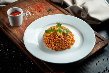 Appetizing and healthy side dish - bulgur in red sauce with Turkish spices in a white plate on a dark background