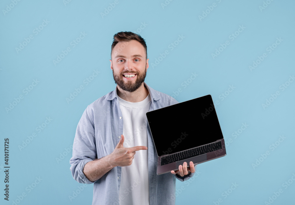 Poster positive young man pointing at empty laptop screen with space for your website design over blue back