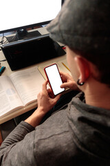 Hombre joven caucásico usando el telefono móvil enfrente de un ordenador y escriorio con libros estudiando a distancia desde casa.