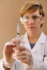 Female nurse doctor wear white coat, protective gloves and glasses holding vaccine syringe against pandemic covid, intended to provide acquired immunity against covid‑19. Vaccination schedule concept.