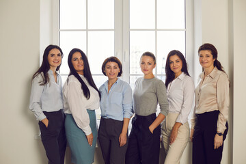 Obraz premium Group portrait of successful confident stylish women in their 20s and 30s standing together by office window. Professional team of happy young company workers or business partners smiling at camera