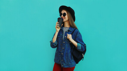 Portrait of young woman drinking a juice wearing a black round hat, denim jacket on a blue background