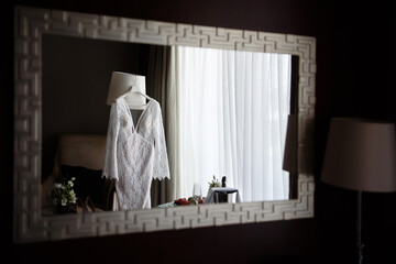 Reflection of beautiful beige wedding dress in mirror in hotel room, copy space. Bride shoes and earings standing on sofa