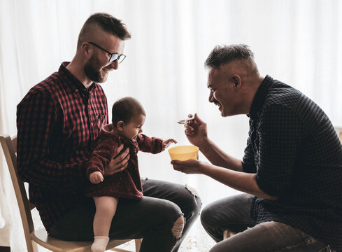 Male gay couple with adopted baby girl at home - Two handsome dads feed the baby girl on kitchen - Male babysitters - Lgbt family at home - Diversity concept
