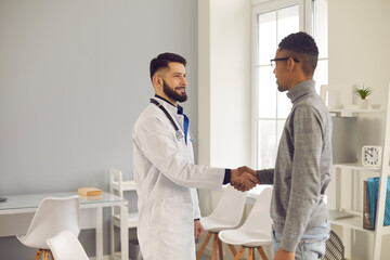 Obraz premium Doctor shaking hands with African-American man in office at clinic or hospital. Smiling young practitioner welcoming new patient. Medical specialist greeting client before consultation or interview