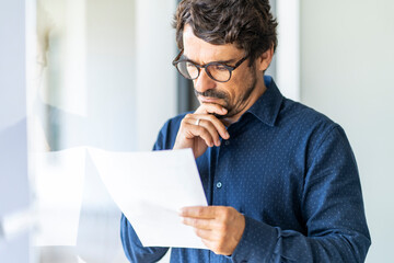 Business man wearing glasses  holding paper document. Successful male portrait thinking and reading contract at the office by the window - Powered by Adobe