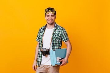 Tourist guy wearing plaid shirt and white T-shirt looking at camera. Portrait of man with diving mask on his head, holding retro camera and hand suitcase
