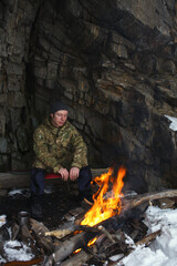 Tired traveller warms up near a campfire divorced next to an overhanging cliff protecting him from the wind.